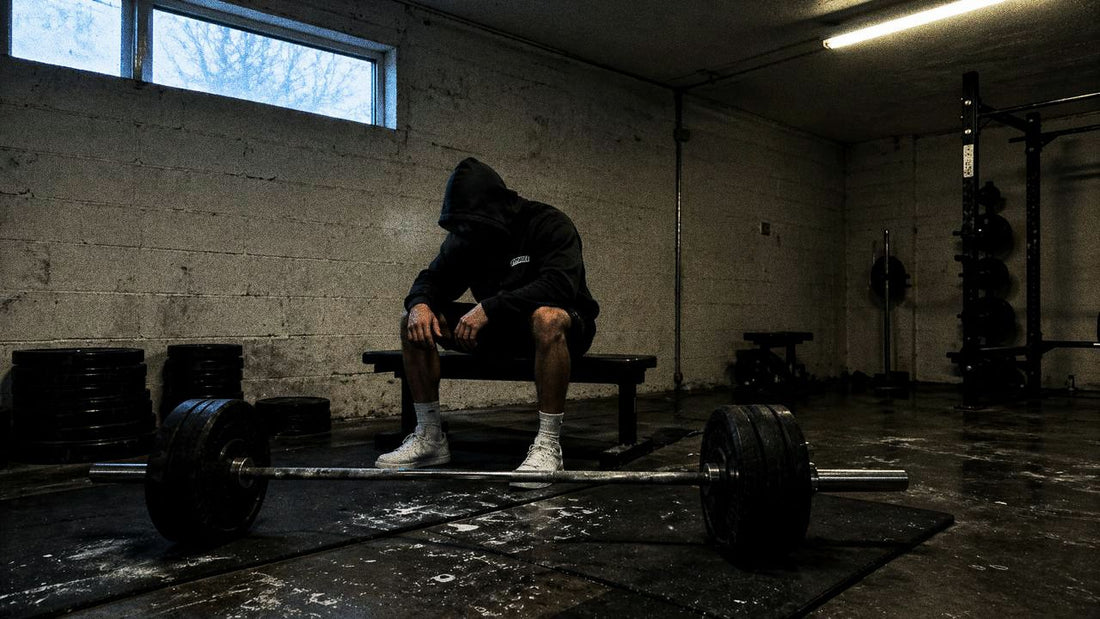 Hood-up lifter sitting on a bench at dawn, cold blue-grey gym light, loaded barbell in foreground.