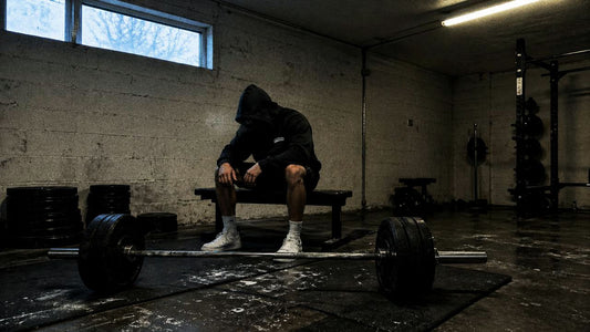 Hood-up lifter sitting on a bench at dawn, cold blue-grey gym light, loaded barbell in foreground.