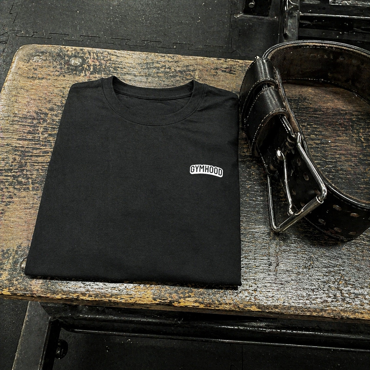 Black GYMHOOD t-shirt folded on a worn wooden gym bench beside a leather lifting belt