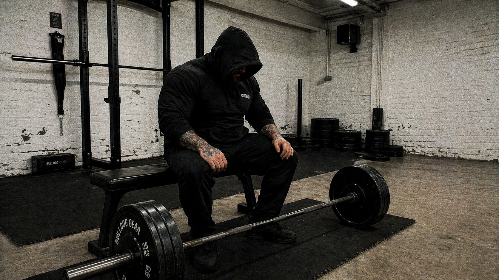 Heavy strength GYMHOOD lifter resting between sets, hood up, beard visible