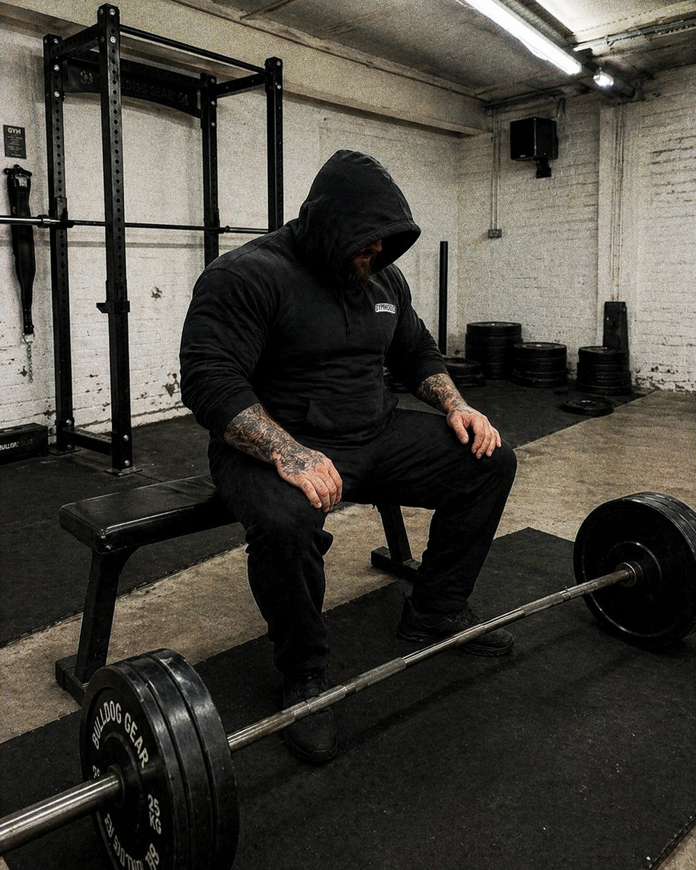 Heavy strength GYMHOOD lifter resting between sets, hood up, beard visible